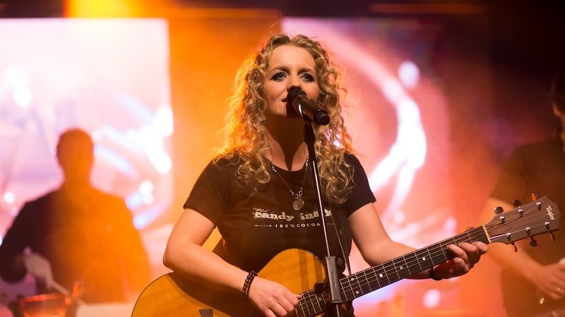 Frau mit Gitarre singt auf der Bühne, umgeben von bunten Lichtern und Nebel., © Elisapark Media GmbH