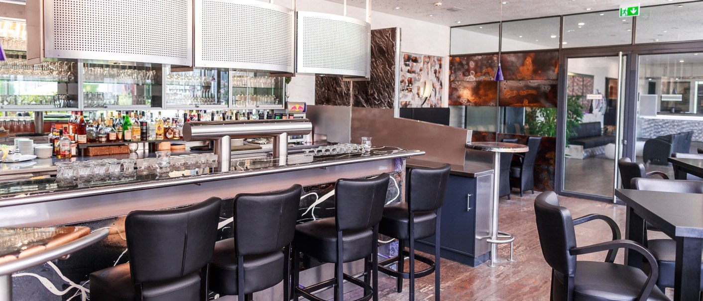 Moderne Bar mit schwarzen Barhockern, Glasregalen und Flaschen im Hintergrund. Der Raum wirkt stilvoll und einladend., © PLAZA Hotelgroup GmbH Moderne Bar mit schwarzen Barhockern, Glasregalen und Flaschen im Hintergrund. Der Raum wirkt stilvoll und einladend., © PLAZA Hotelgroup GmbH