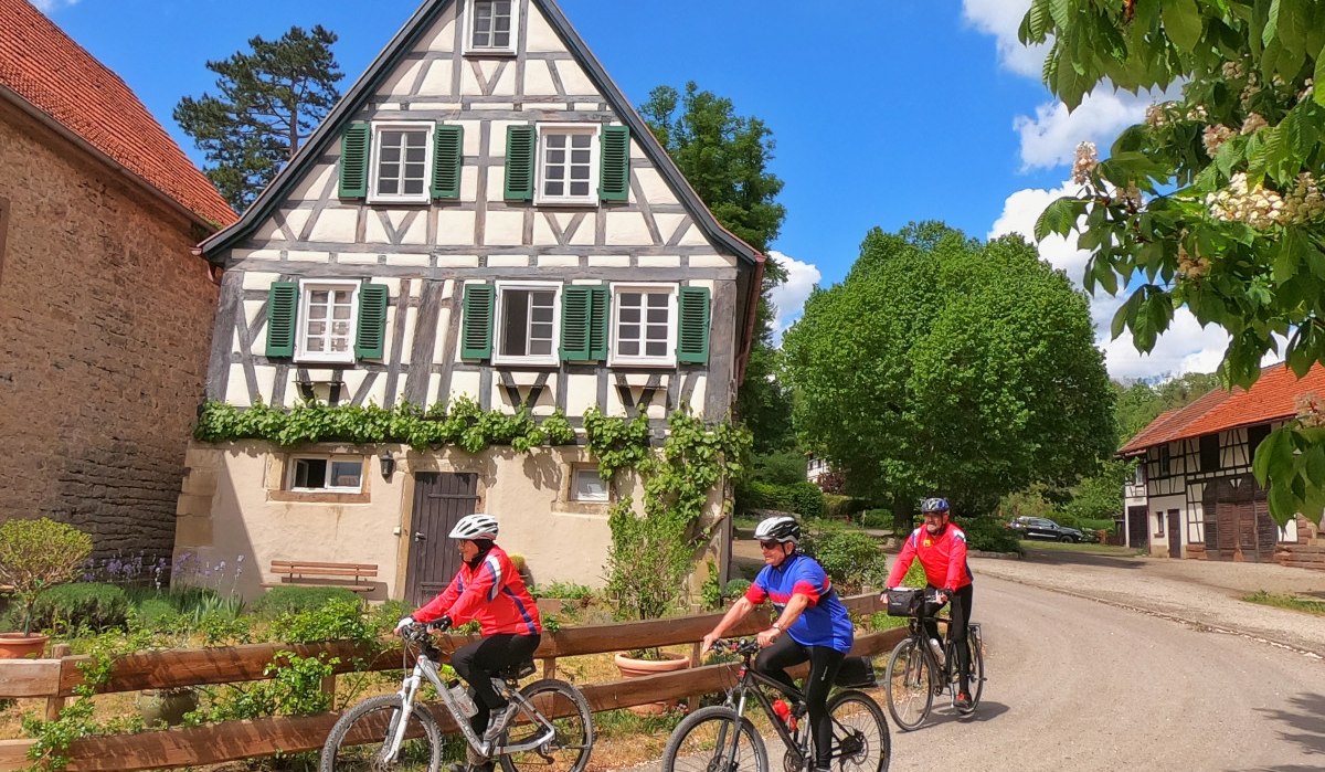 Drei Radfahrer in bunter Kleidung fahren an einem malerischen Fachwerkhaus vorbei, umgeben von grünen Bäumen und blauem Himmel., © Land der 1000 Hügel - Kraichgau-Stromberg Drei Radfahrer in bunter Kleidung fahren an einem malerischen Fachwerkhaus vorbei, umgeben von grünen Bäumen und blauem Himmel., © Land der 1000 Hügel - Kraichgau-Stromberg
