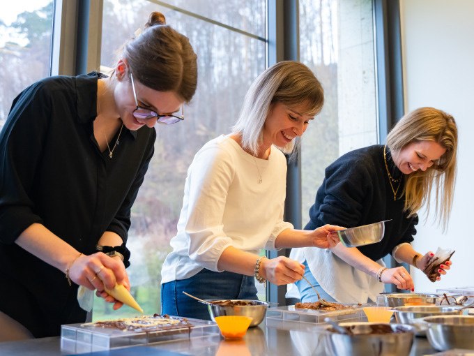 Drei Frauen dekorieren Schokolade in einer Werkstatt. Sie lachen und verwenden verschiedene Werkzeuge, um die Schokolade zu gestalten., &copy; Alfred Ritter GmbH &amp; Co. KG | Waldenbuch