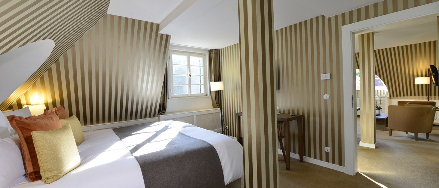 Elegante Hotelsuite mit gestreiften Wänden, gemütlichem Bett und angrenzendem Wohnzimmer. Helle Fenster und stilvolle Einrichtung schaffen eine einladende Atmosphäre., © Waldhotel Stuttgart GmbH Elegante Hotelsuite mit gestreiften Wänden, gemütlichem Bett und angrenzendem Wohnzimmer. Helle Fenster und stilvolle Einrichtung schaffen eine einladende Atmosphäre., © Waldhotel Stuttgart GmbH