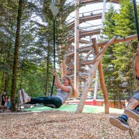 Zwei Kinder schaukeln fröhlich im Abenteuerwald Sommerberg. Im Hintergrund ist ein großer Holzpfad zu sehen, umgeben von hohen Bäumen., © Erlebnis Akademie AG
