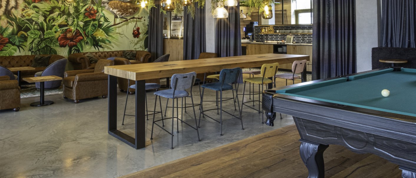 Gemütliche Lounge mit Billardtisch, Holztisch und bunten Stühlen. Im Hintergrund eine Wand mit Pflanzenmotiv und eine Bar., © 13 6 Design GmbH (Judenhof 10, 89073 Ulm)
