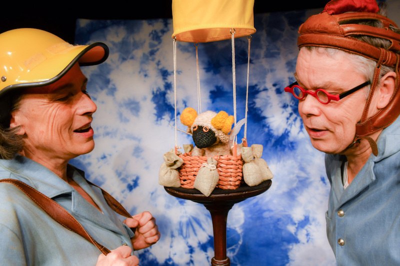 Zwei Personen mit Helmen betrachten ein gestricktes Schaf in einem Mini-Heißluftballonkorb vor blau-weißem Hintergrund., © FILharmonie Filderstadt Zwei Personen mit Helmen betrachten ein gestricktes Schaf in einem Mini-Heißluftballonkorb vor blau-weißem Hintergrund., © FILharmonie Filderstadt