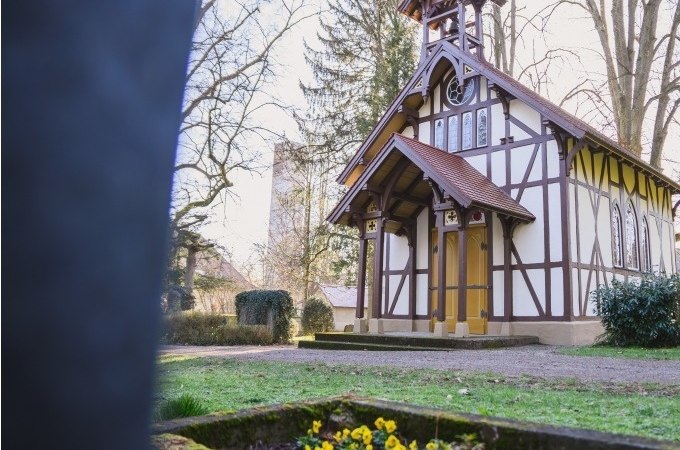 Eine malerische Fachwerkkapelle auf einem Friedhof, umgeben von Bäumen. Im Vordergrund blühen gelbe Blumen in einem Grab., © Eigenbetrieb Tourismus und Citymanagement Schorndorf Eine malerische Fachwerkkapelle auf einem Friedhof, umgeben von Bäumen. Im Vordergrund blühen gelbe Blumen in einem Grab., © Eigenbetrieb Tourismus und Citymanagement Schorndorf