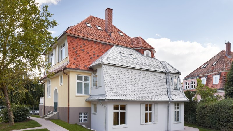 Zweistöckiges Haus mit rotem Ziegeldach, gelber Fassade und weißen Fenstern, umgeben von Bäumen und Rasen., © Philip Kottlorz