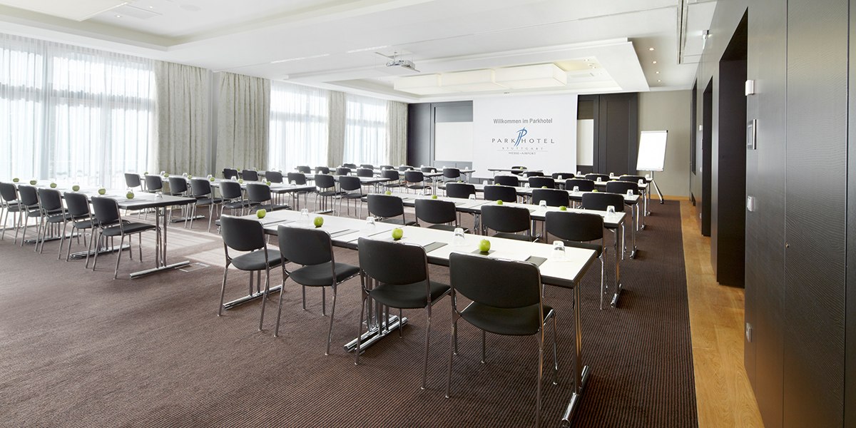 Ein moderner Konferenzraum mit Reihen von Tischen und Stühlen, einer Leinwand und einem Flipchart. Helle Vorhänge lassen Tageslicht herein., © Parkhotel Stuttgart Messe-Airport