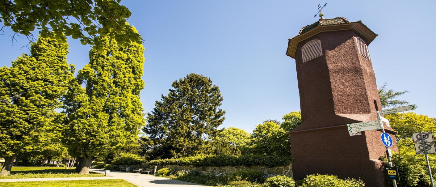 Ein roter Turm am Alten Friedhof in Fellbach, umgeben von hohen Bäumen und Wegweisern, bei klarem, sonnigem Himmel., © SMG, Sarah Schmid
