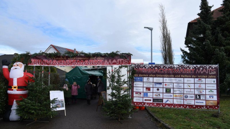 Eingang zum Weihnachtsmarkt Maitis mit einem gro&szlig;en Weihnachtsmann, Tannenb&auml;umen und einem Banner mit Unterst&uuml;tzerlogos.