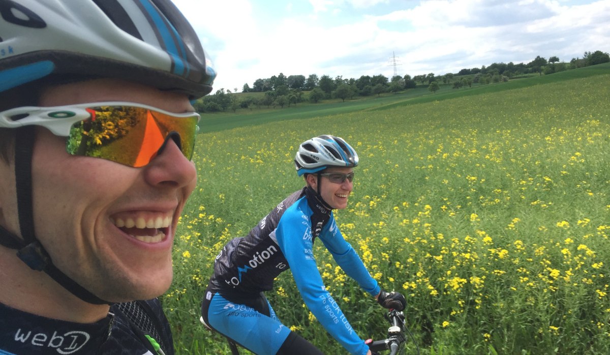 Zwei Fahrradfahrer in blauer Sportkleidung und Helmen fahren lächelnd durch ein gelbes Blumenfeld unter bewölktem Himmel., © www.pro-cycl.de Zwei Fahrradfahrer in blauer Sportkleidung und Helmen fahren lächelnd durch ein gelbes Blumenfeld unter bewölktem Himmel., © www.pro-cycl.de