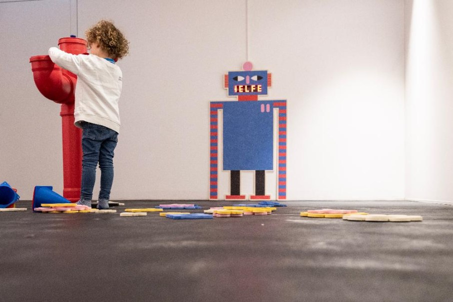 Ein Kind spielt in einem Raum mit einem roten Objekt. Auf der Wand ist ein Roboterbild mit der Aufschrift 'Helfe'. Bunte Scheiben liegen auf dem Boden., &copy; Ludwigsburg Museum