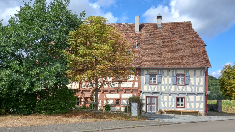 Zweigeteiltes Fachwerkhaus - linke Haushälfte mit rotem Fachwerk und hellesn Gefachen, rechte Haushälfte mit hellgrauem Fachwerk. Das Gebäude hat ein rotes Ziegeldach und ist von Bäumen umgeben., © Petra Natzkowski Zweigeteiltes Fachwerkhaus - linke Haushälfte mit rotem Fachwerk und hellesn Gefachen, rechte Haushälfte mit hellgrauem Fachwerk. Das Gebäude hat ein rotes Ziegeldach und ist von Bäumen umgeben., © Petra Natzkowski
