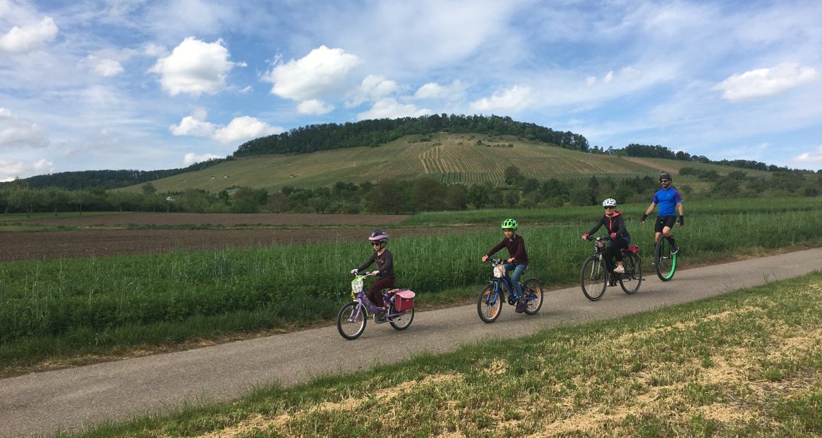 Eine Familie fährt auf einem Radweg durch eine ländliche Landschaft. Im Hintergrund ist ein bewachsener Hügel unter blauem Himmel zu sehen., © Land der 1000 Hügel - Kraichgau-Stromberg Eine Familie fährt auf einem Radweg durch eine ländliche Landschaft. Im Hintergrund ist ein bewachsener Hügel unter blauem Himmel zu sehen., © Land der 1000 Hügel - Kraichgau-Stromberg