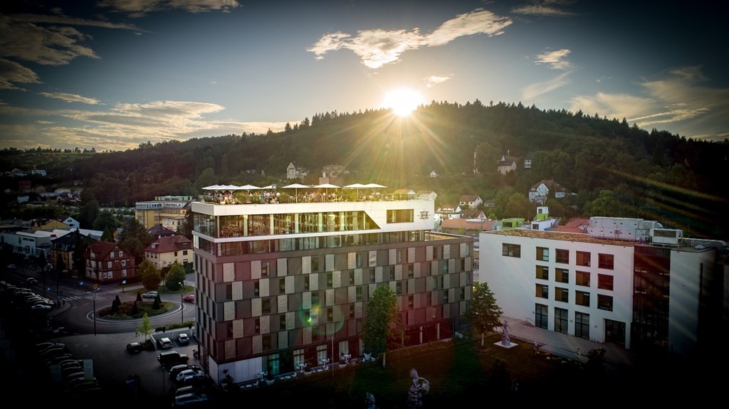 Modernes Hotelgebäude bei Sonnenuntergang, umgeben von grüner Landschaft und Häusern. Die Sonne strahlt über einem bewaldeten Hügel., © Hotel am Remspark Modernes Hotelgebäude bei Sonnenuntergang, umgeben von grüner Landschaft und Häusern. Die Sonne strahlt über einem bewaldeten Hügel., © Hotel am Remspark