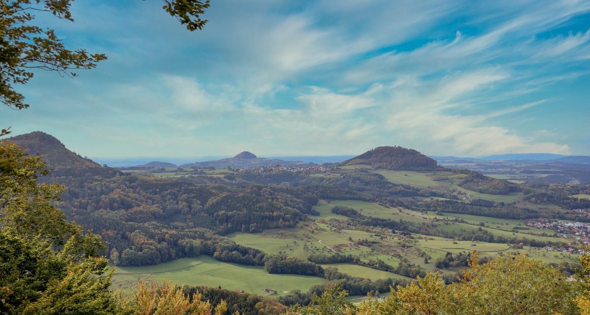 Drei bewaldete Berge in weiter ferne. Davor liegen Wiesen, Bäume und Wälder. Alles ist weit entfernt von oben betrachtet und deshalb sehr klein. Der Himmel ist blau mit weisen Schleierwolken., © Foto: Mario Klaiber Drei bewaldete Berge in weiter ferne. Davor liegen Wiesen, Bäume und Wälder. Alles ist weit entfernt von oben betrachtet und deshalb sehr klein. Der Himmel ist blau mit weisen Schleierwolken., © Foto: Mario Klaiber