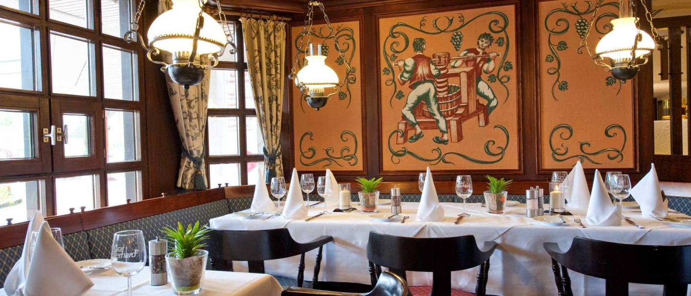 Elegantes Restaurant mit gedecktem Tisch, weißen Servietten, Wandmalerei von Weinbauern und Kronleuchtern. Gemütliche Atmosphäre mit Holzelementen., © Biosphärenhotel Graf Eberhard