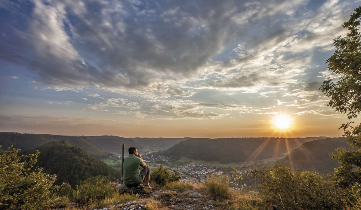 Eine Person sitzt auf einem Felsen und blickt auf ein Tal bei Sonnenuntergang. Der Himmel ist teils bewölkt, die Sonne strahlt über die Landschaft., © Landkreis Göppingen Eine Person sitzt auf einem Felsen und blickt auf ein Tal bei Sonnenuntergang. Der Himmel ist teils bewölkt, die Sonne strahlt über die Landschaft., © Landkreis Göppingen