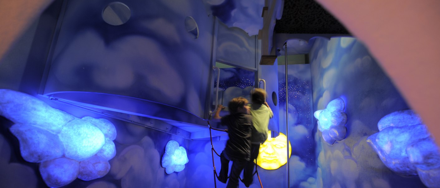 Zwei Kinder klettern in einem blau beleuchteten Raum mit Wolken und einem leuchtenden Mond an der Wand., © Sensapolis Sindelfingen