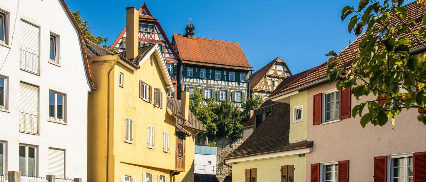 Blick auf bunte Fachwerkhäuser in einer Altstadt unter klarem, blauem Himmel. Eine Person geht die Straße entlang., © Stuttgart-Marketing GmbH, Sarah Schmid Blick auf bunte Fachwerkhäuser in einer Altstadt unter klarem, blauem Himmel. Eine Person geht die Straße entlang., © Stuttgart-Marketing GmbH, Sarah Schmid