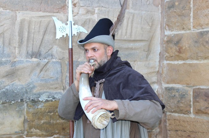 Ein Mann in historischer Kleidung bläst ein Horn vor einer alten Steinmauer. Er trägt einen Hut mit Feder und hält eine Hellebarde., © Touristik und Marketing GmbH Ein Mann in historischer Kleidung bläst ein Horn vor einer alten Steinmauer. Er trägt einen Hut mit Feder und hält eine Hellebarde., © Touristik und Marketing GmbH