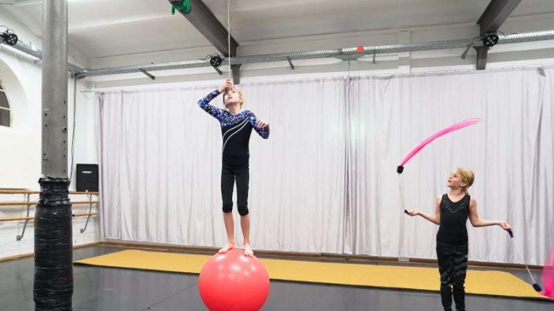 Zwei Kinder üben Zirkuskünste: Eines balanciert auf einem roten Ball, das andere schwingt ein pinkes Band. Der Raum hat hohe Decken und Vorhänge., © Kunstschule Labyrinth