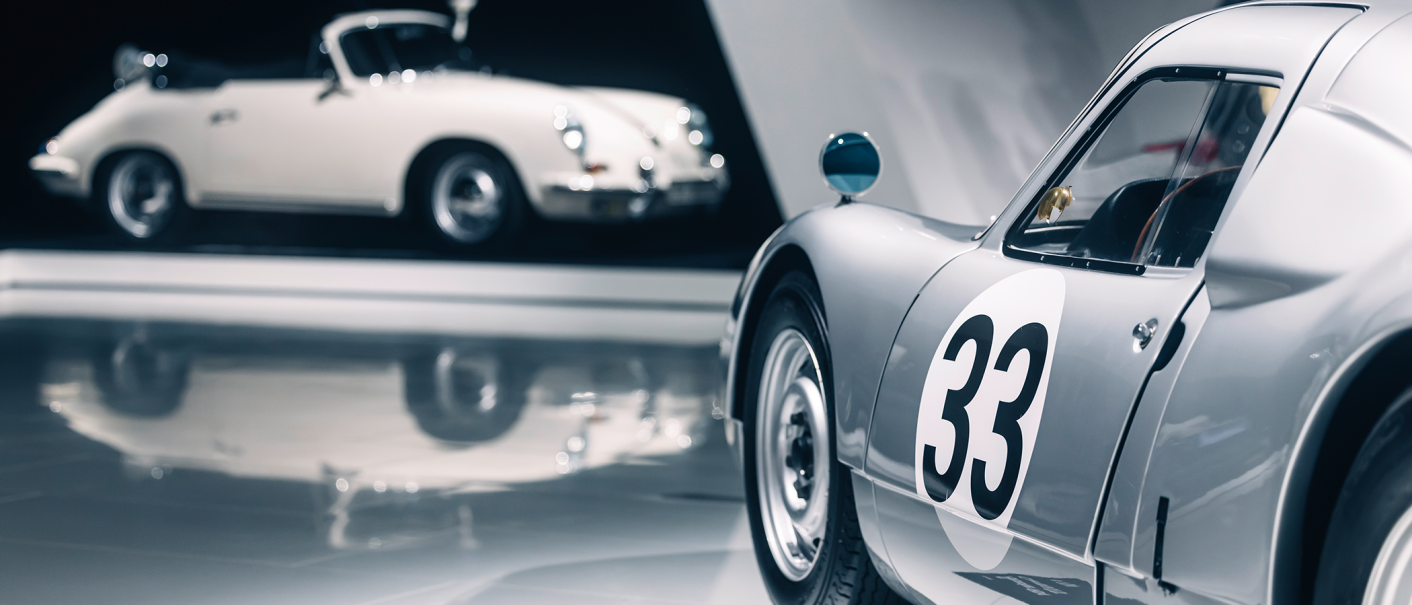 Zwei klassische Autos in einer Ausstellung. Ein weißes Cabrio mit Polizeilicht und ein silbernes Auto mit der Nummer 33 im Vordergrund., © Porsche AG Zwei klassische Autos in einer Ausstellung. Ein weißes Cabrio mit Polizeilicht und ein silbernes Auto mit der Nummer 33 im Vordergrund., © Porsche AG