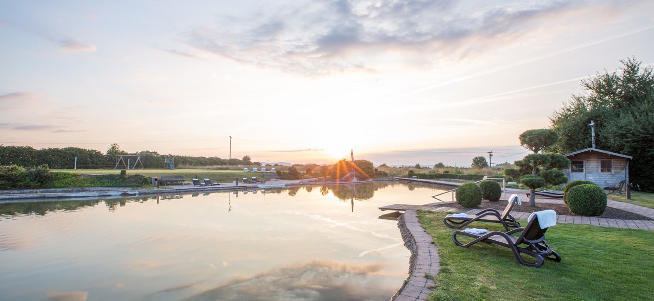 Naturbadesee im ARAMIS Hotel bei Sonnenuntergang, umgeben von Liegestühlen und gepflegtem Garten., © ARAMIS Hotel & Freizeit GmbH