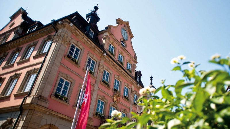 Ein historisches Gebäude mit Uhr und Flaggen, umgeben von blühenden Pflanzen, unter einem klaren blauen Himmel., © LGS Schwäbisch Gmünd