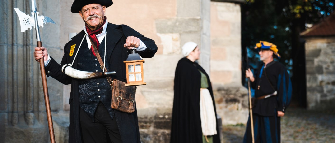 Ein Mann in historischer Kleidung mit Laterne und Hellebarde. Zwei weitere Personen in mittelalterlicher Kleidung im Hintergrund., © Stadt Nürtingen, U. Jendrass