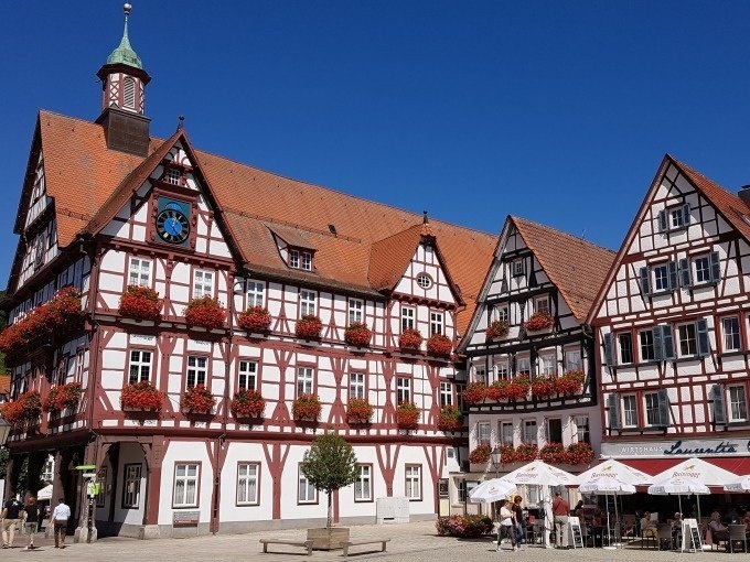 Fachwerkh&auml;user in Bad Urach mit roten Blumen geschm&uuml;ckt, blauer Himmel. Menschen sitzen unter Sonnenschirmen vor einem Caf&eacute;., &copy; SwabianTravel