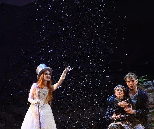 Drei Schauspieler auf der Bühne in einer Szene aus Peer Gynt. Eine Frau im weißen Kleid mit Zylinder steht, während zwei Personen auf einem Felsen sitzen. Schneefall., © Backnang Drei Schauspieler auf der Bühne in einer Szene aus Peer Gynt. Eine Frau im weißen Kleid mit Zylinder steht, während zwei Personen auf einem Felsen sitzen. Schneefall., © Backnang