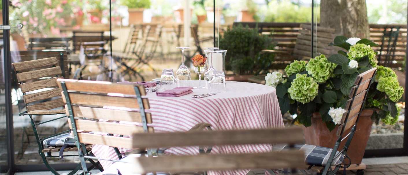 Ein einladend gedeckter Tisch mit rot-wei&szlig; gestreifter Tischdecke in einem Gartenrestaurant. Umgeben von Holzm&ouml;beln und bl&uuml;henden Pflanzen., &copy; Valentino