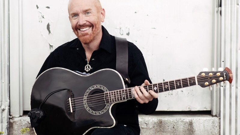 Ein Mann mit rotem Bart sitzt lächelnd auf einer Treppe und hält eine schwarze Akustikgitarre. Der Hintergrund ist eine weiße Wand., © Mauerwerk GmbH Ein Mann mit rotem Bart sitzt lächelnd auf einer Treppe und hält eine schwarze Akustikgitarre. Der Hintergrund ist eine weiße Wand., © Mauerwerk GmbH
