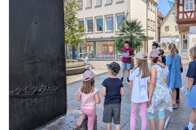 Kindergruppe hört einem jungen Mann in einer Stadt zu. Im Hintergrund sind Gebäude und ein Denkmal sichtbar., © Eigenbetrieb Tourismus und Citymanagement Schorndorf