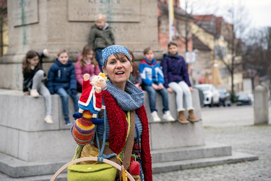 Eine Frau mit bunter Kleidung und Handpuppe steht vor einer Gruppe von Kindern, die auf einem Denkmal sitzen., &copy; Tourist Information und Ticket Service Ludwigsburg