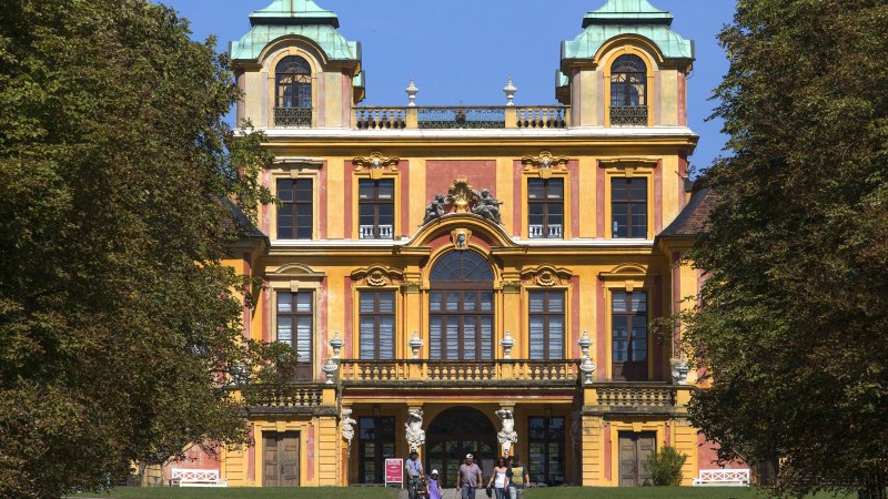 Barockes Schloss mit gelber Fassade und grünen Dächern, umgeben von Bäumen. Menschen stehen vor dem Eingang., © Tourismus & Events Ludwigsburg Barockes Schloss mit gelber Fassade und grünen Dächern, umgeben von Bäumen. Menschen stehen vor dem Eingang., © Tourismus & Events Ludwigsburg