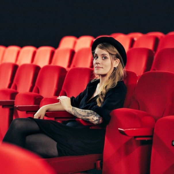 Eine Person mit Hut und tätowiertem Arm sitzt entspannt in einem leeren Kinosaal auf einem roten Sitz., © Mauerwerk GmbH Eine Person mit Hut und tätowiertem Arm sitzt entspannt in einem leeren Kinosaal auf einem roten Sitz., © Mauerwerk GmbH