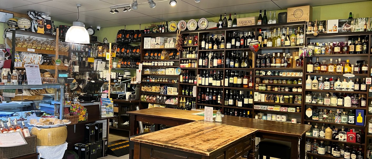 Feinkostladen mit Regalen voller Wein, Spirituosen und Delikatessen. Ein Holztisch mit St&uuml;hlen steht im Vordergrund. Der Raum ist hell beleuchtet., &copy; Feinkost-Galeria