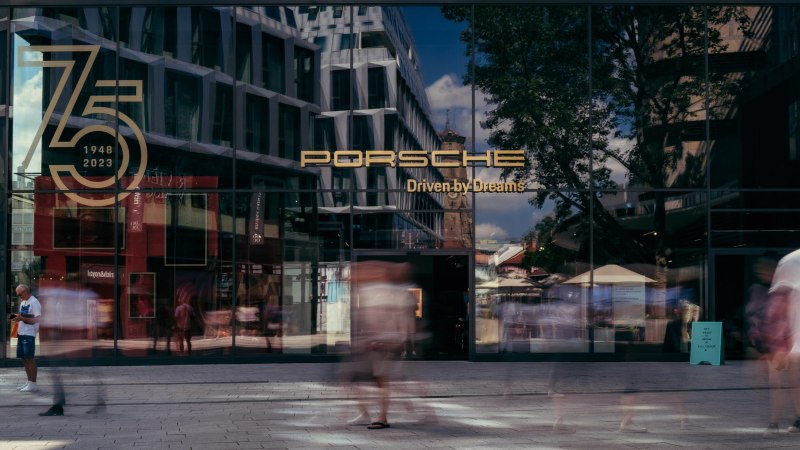 Glasfassade des Porsche Brand Stores mit Jubiläumslogo '75 Jahre 1948-2023'. Menschen gehen vorbei, Gebäude und Bäume spiegeln sich im Glas., © Porsche AG