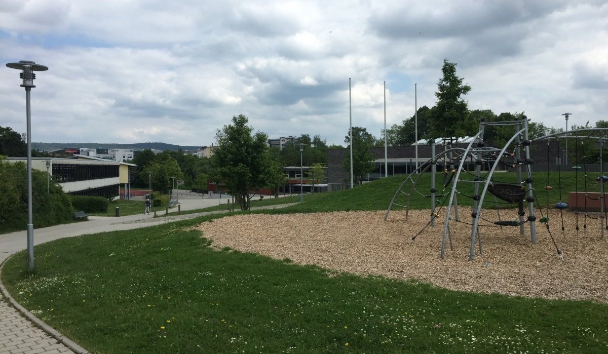 Ein moderner Spielplatz mit Kletterger&uuml;st auf einer Gr&uuml;nfl&auml;che, umgeben von Geb&auml;uden und Gehwegen unter bew&ouml;lktem Himmel., &copy; Natur.Nah. Sch&ouml;nbuch & Heckeng&auml;u