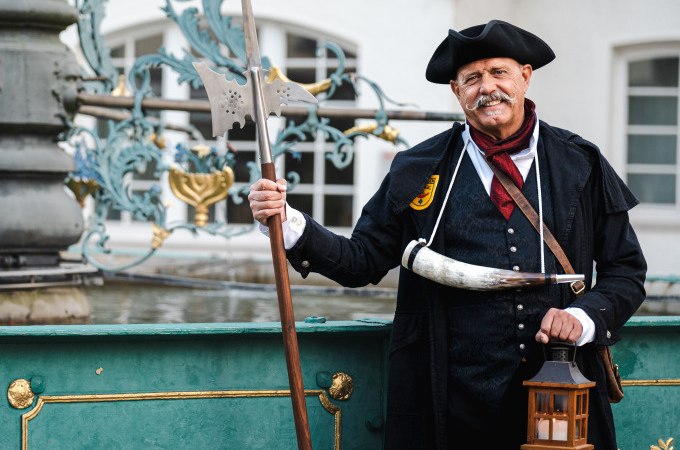 Mann in historischer Nachtwächterkleidung mit Hellebarde und Laterne vor einem verzierten Brunnen., © Stadt Nürtingen