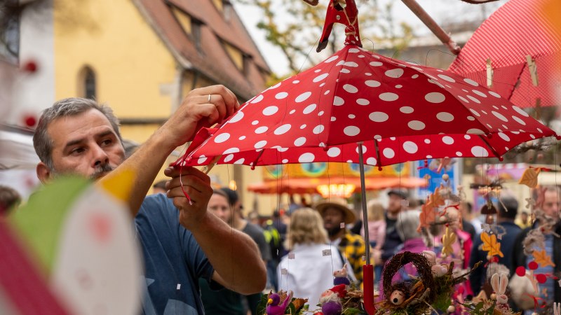 Ein Mann dekoriert einen roten Regenschirm mit weißen Punkten auf einem Kunsthandwerkermarkt. Im Hintergrund sind Besucher und bunte Stände zu sehen., © Stadt Nürtingen Ein Mann dekoriert einen roten Regenschirm mit weißen Punkten auf einem Kunsthandwerkermarkt. Im Hintergrund sind Besucher und bunte Stände zu sehen., © Stadt Nürtingen