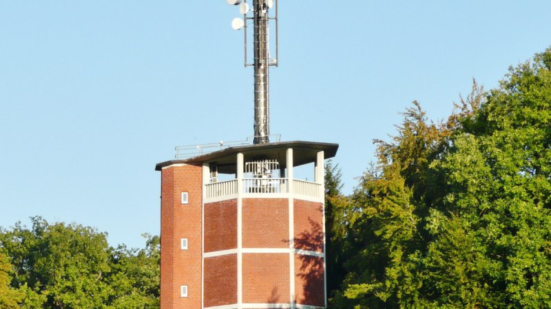 Roter Backstein-Wasserturm mit Antennenmast, umgeben von gr&uuml;nen B&auml;umen unter klarem Himmel.