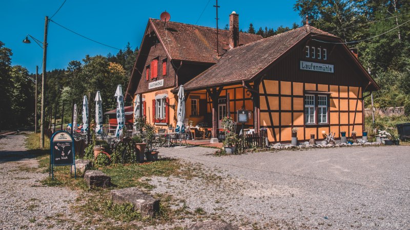 Fachwerkgeb&auml;ude 'Laufenm&uuml;hle' mit Sonnenschirmen und Au&szlig;enbereich, umgeben von Wald, unter blauem Himmel., &copy; Stadt Welzheim