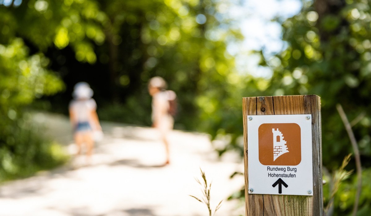 Ein Wegweiser mit der Aufschrift 'Rundweg Burg Hohenstaufen' zeigt den Weg. Im Hintergrund sind zwei unscharfe Personen auf einem Waldweg., © TMBW