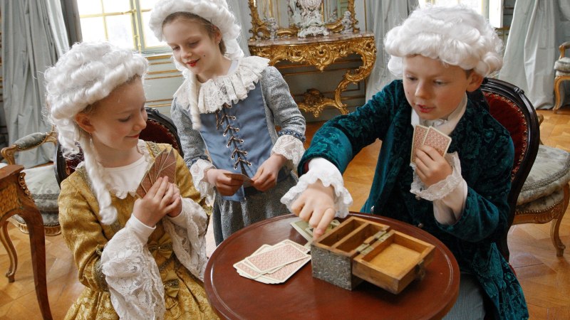 Drei Kinder in historischen Kostümen spielen Karten in einem prunkvollen Raum mit antiken Möbeln und Perücken., © Tourismus und Events Ludwigsburg