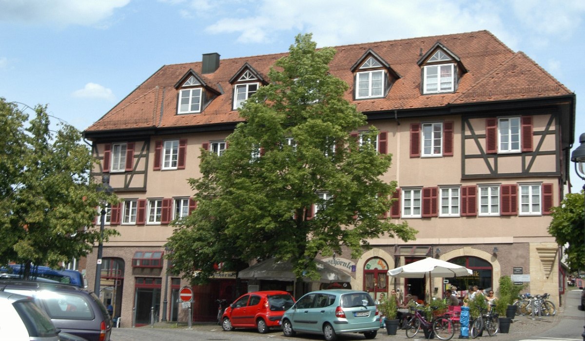 Fachwerkhaus mit roten Fensterläden, geparkte Autos und Fahrräder. Menschen sitzen unter Sonnenschirmen vor einem Café. Bäume im Vordergrund., © Natur.Nah. Schönbuch & Heckengäu Fachwerkhaus mit roten Fensterläden, geparkte Autos und Fahrräder. Menschen sitzen unter Sonnenschirmen vor einem Café. Bäume im Vordergrund., © Natur.Nah. Schönbuch & Heckengäu