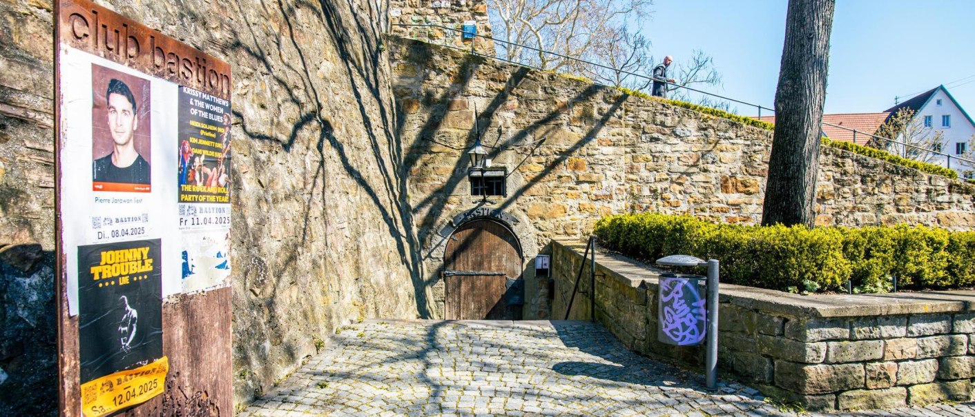 Eingang zur Club Bastion in Kirchheim unter Teck, umgeben von alten Steinmauern. Plakate für Veranstaltungen sind an der Wand angebracht., © Stuttgart-Marketing GmbH, Sarah Schmid