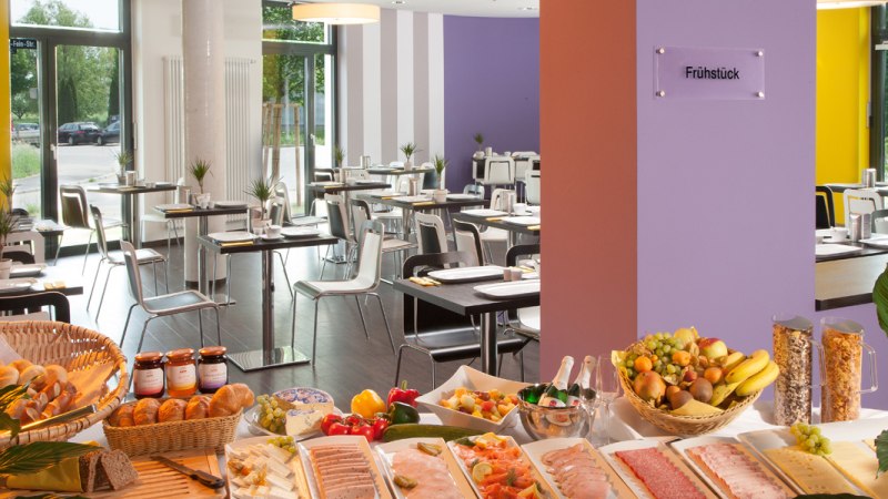 Ein reichhaltiges Frühstücksbuffet mit Brot, Aufschnitt, Obst und Müsli in einem modernen Restaurant mit bunten Wänden., © Star Inn Stuttgart Airport-Messe