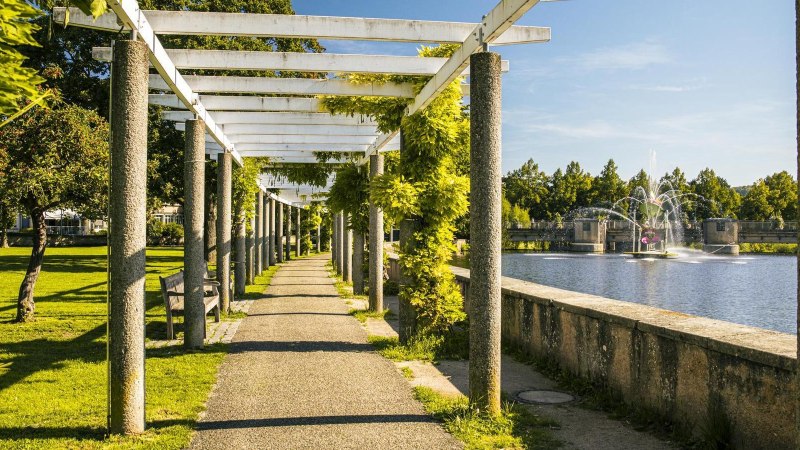 Ein sonniger Weg mit Pergola und Bänken führt entlang eines künstlichen Sees mit Springbrunnen. Grüne Bäume und Pflanzen säumen den Weg., © Stuttgart-Marketing GmbH, Sarah Schmid